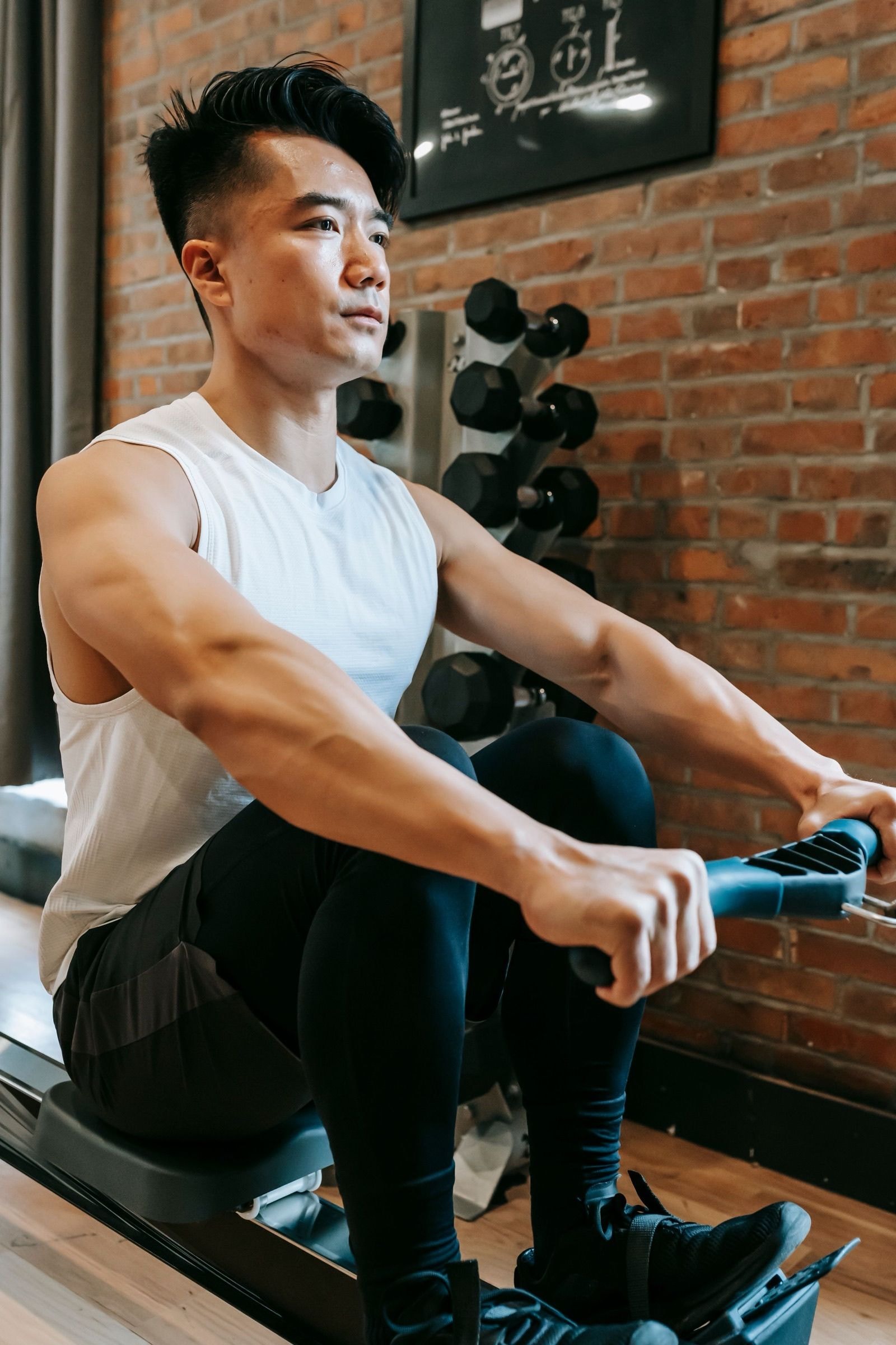 A man in a white tank top pulling on an indoor rower beside a brick wall